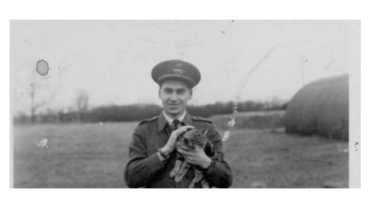 Smiling man in a military uniform holding a small cat outdoors in a field.