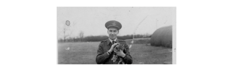 Smiling man in a military uniform holding a small cat outdoors in a field.