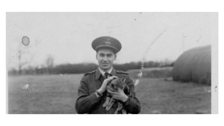 Smiling man in a military uniform holding a small cat outdoors in a field.