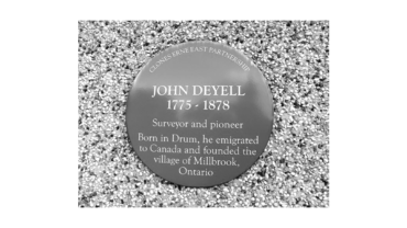 Circular commemorative plaque on gravel: 'John Deyell 1775–1878' with text about a surveyor and pioneer and the village of Millbrook, Ontario.