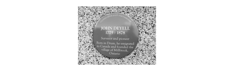 Circular commemorative plaque on gravel: 'John Deyell 1775–1878' with text about a surveyor and pioneer and the village of Millbrook, Ontario.