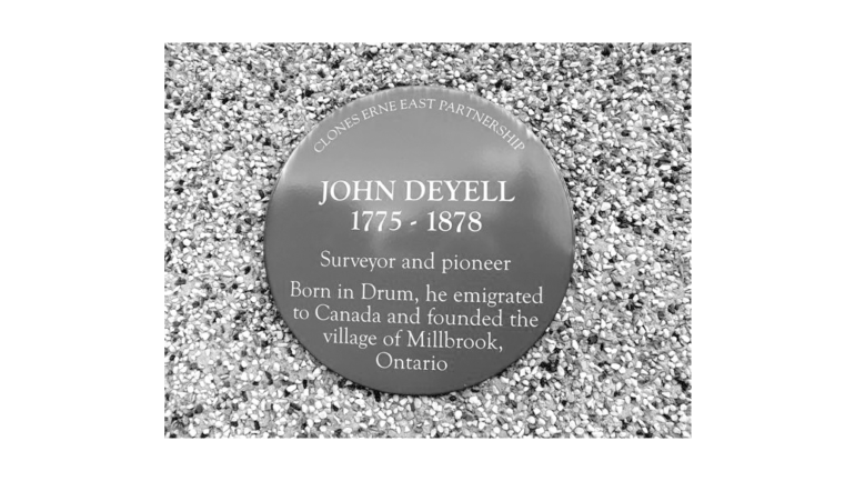 Circular commemorative plaque on gravel: 'John Deyell 1775–1878' with text about a surveyor and pioneer and the village of Millbrook, Ontario.