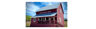 Red two-story mill building labeled 'Needler's Mill' with a white-trimmed porch and metal roof on a sunny day, blue sky above.