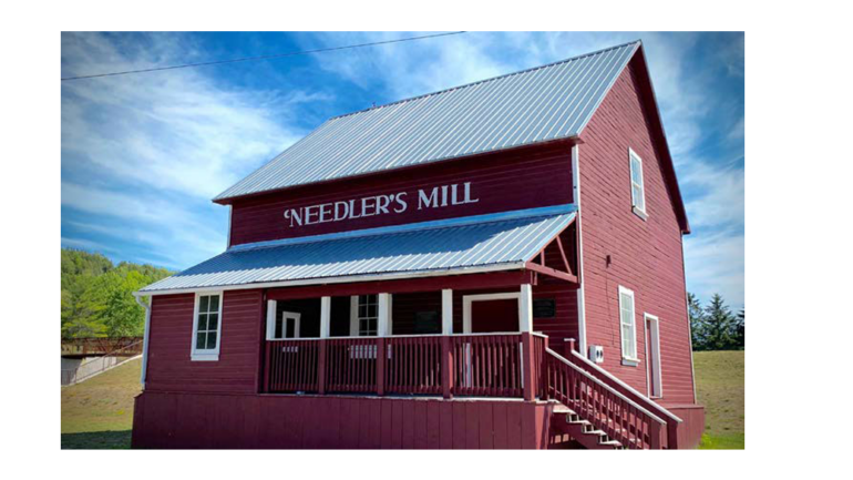 Red two-story mill building labeled 'Needler's Mill' with a white-trimmed porch and metal roof on a sunny day, blue sky above.