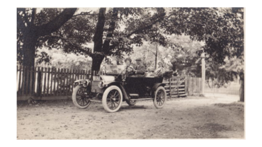 Vintage open-top car with four passengers on a dirt road beside a wooden fence under trees in a black-and-white photo.
