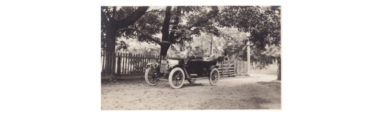 Vintage open-top car with four passengers on a dirt road beside a wooden fence under trees in a black-and-white photo.