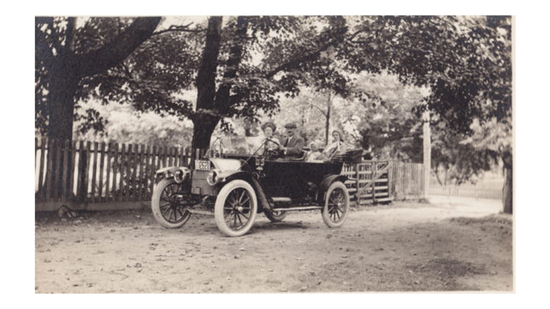 Vintage open-top car with four passengers on a dirt road beside a wooden fence under trees in a black-and-white photo.