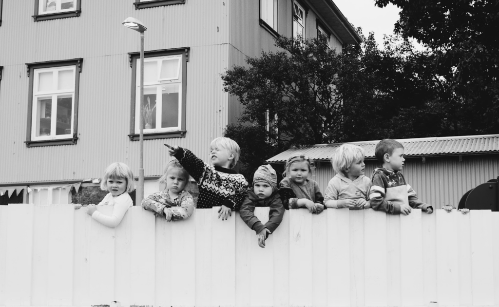 Children lean over a white fence, peering and pointing at something off to the right in a residential area (black-and-white photo).