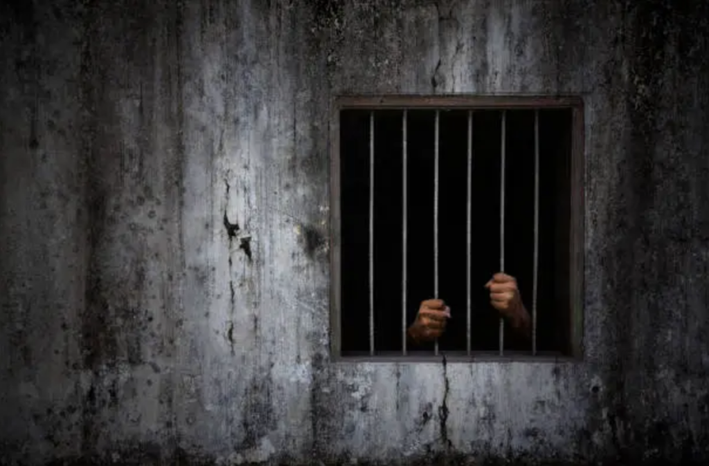 Hands gripping vertical prison bars through a small window, set in a cracked, grimy concrete wall in a dark cell.