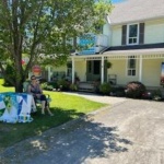 Two people sit at a blue-and-white table on the lawn in front of a beige two-story house, with a driveway and a sign/banner visible in the window.