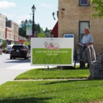 Street scene with a green event sign that reads 'Stop on the tour here! 10am-4pm' and floral graphics, near a man in a hat sitting by a building.