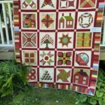 Colorful patchwork quilt hanging over a railing outside, showing many square blocks with red, green, and white geometric patterns.
