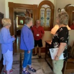 Group of four older adults in a tiled foyer, conversing near a wood-framed doorway and a stained-glass arched window.