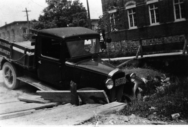 Vintage black pickup truck parked on a wooden dock beside a stone canal, with a brick building in the background.