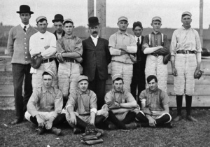 Group of men in vintage baseball uniforms posing outdoors; several wearing caps, one in a suit at the center.