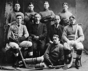 Vintage ice hockey team portrait: eight players in knit sweaters with two coaches posing in a studio with hockey sticks laid across their laps and on the floor.