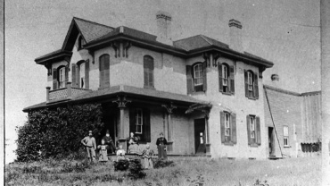 Two-story Victorian-style house with ornate brackets; family stands on the front lawn.