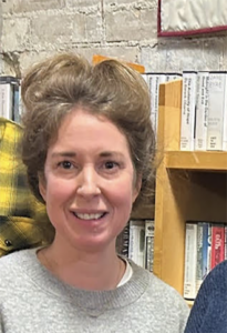 Woman smiling at the camera, wearing a light gray sweater, in front of a bookshelf-filled backdrop in a library or shop.