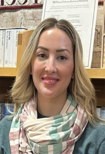 Portrait of a smiling woman with blonde hair wearing a pastel scarf, standing in a bookstore or library with books on shelves behind her.
