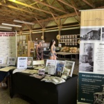 Exhibit booth for a historical society with banners, posters, and tables of pamphlets; two visitors browse the displays.