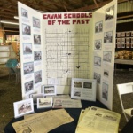 Tri-fold exhibit board titled 'Cavan Schools of the Past' with maps, historical photos, and captions; papers laid out on a black table in front.