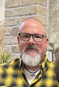 Portrait of a middle-aged man with black-framed glasses and a gray beard, wearing a yellow plaid shirt, indoors beside a brick wall.