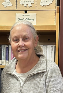Smiling woman with gray hair in a light gray sweater, standing in front of a library shelf labeled 'Seed Library.'