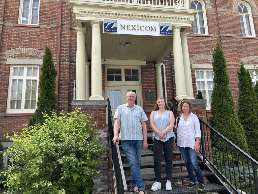 Four people stand on the front steps of a red brick building with a Nexicom sign above the entrance.