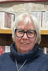 Portrait of an older woman with white hair and black-framed glasses, wearing a blue turtleneck and pearl necklace, in front of a bookshelf.