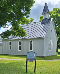 Small gray church with a steep metal roof and steeple, arched windows, and a sign on the lawn in front reading 'The Marsh Trinity Church Anglican'